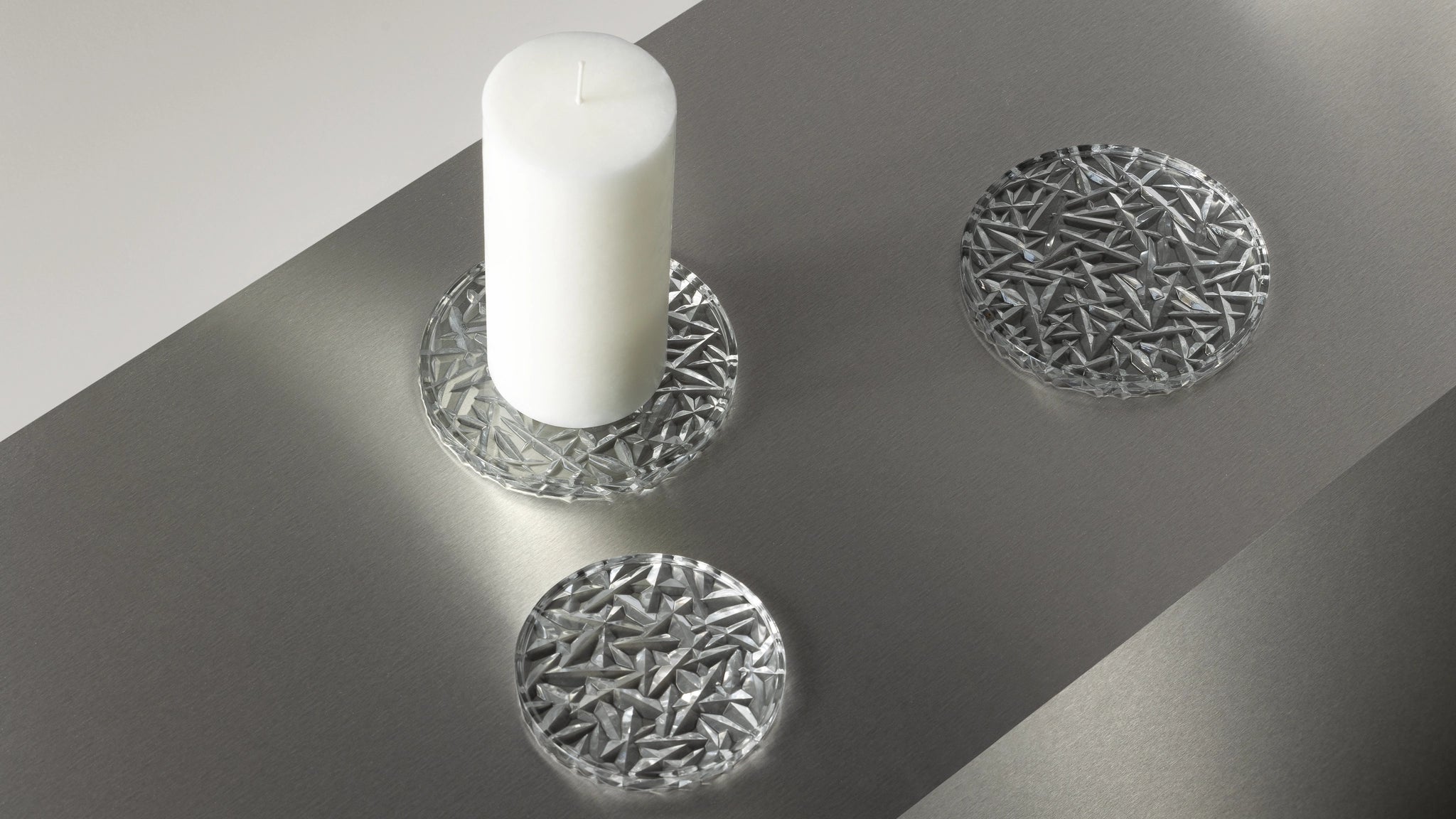 A white candle sits elegantly on an Orrefors: Carat Coaster Small from a 2-Pack, with two coasters resting on a sleek gray surface.