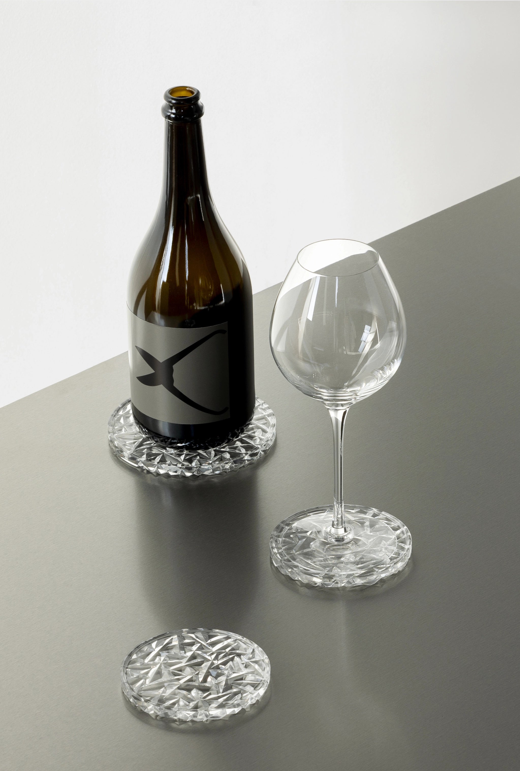 A wine bottle and an empty glass rest elegantly on a gray surface, each supported by a small Orrefors Carat Coaster from the 2-Pack. A third crystal coaster shimmers close by, enhancing the sophisticated tableau.