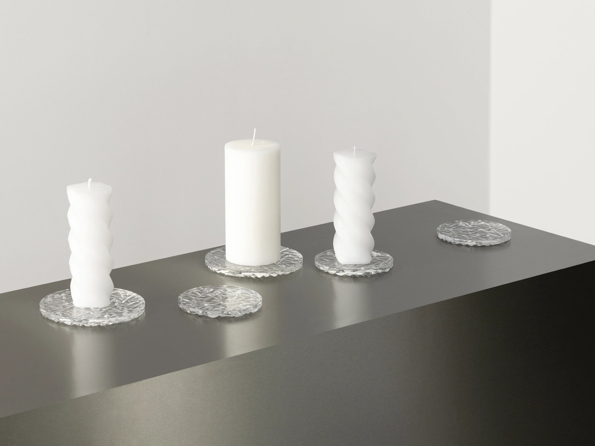 A set of Orrefors Carat Coaster Small (2-Pack) cradles three white pillar candles alongside two textured glass holders on a black surface near a white wall.