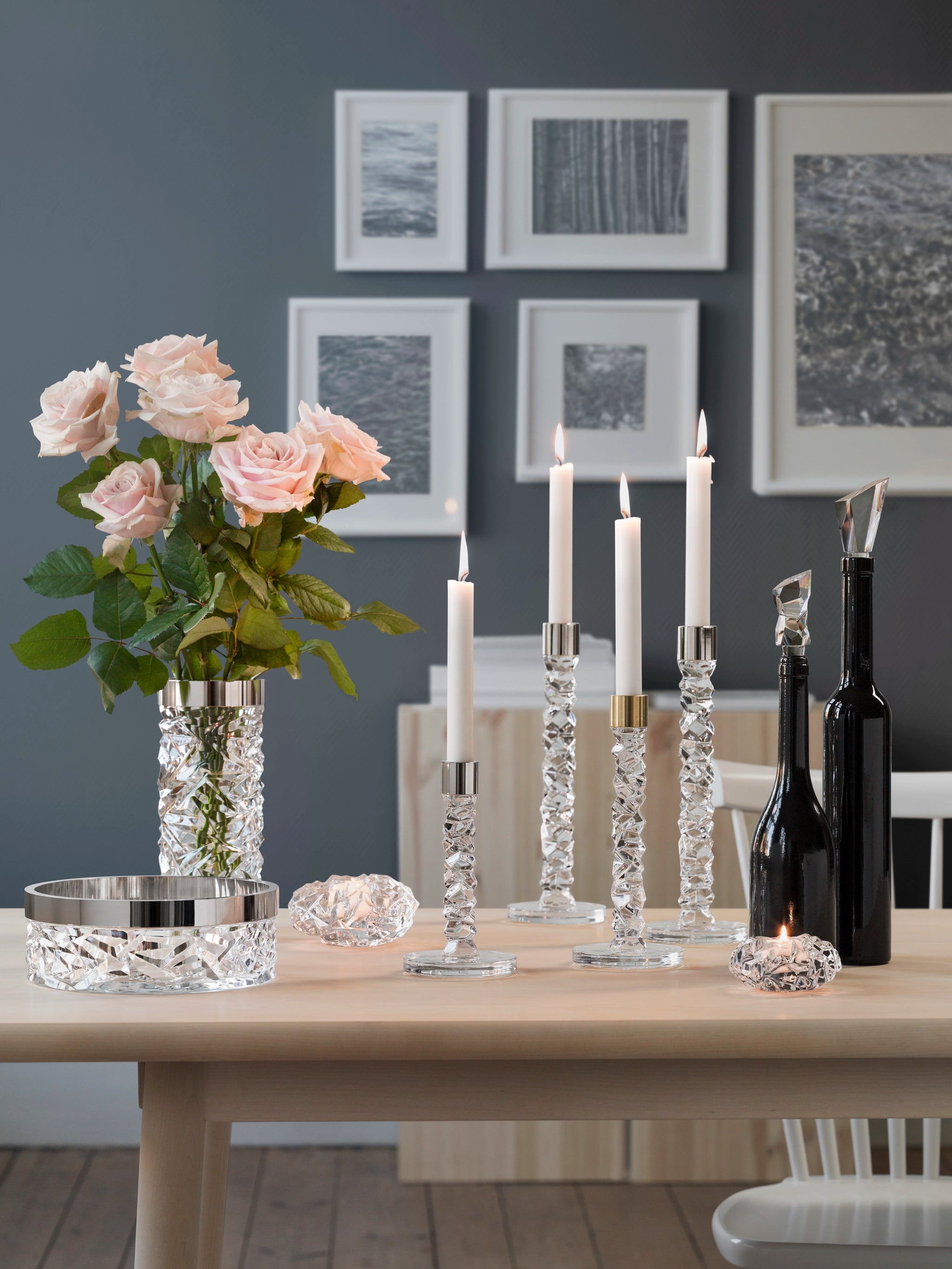 A table displays five lit candles in Orrefors: Carat Votive Large Candle Holders, accompanied by a vase with pink roses, black bottles, and framed wall art in the background.