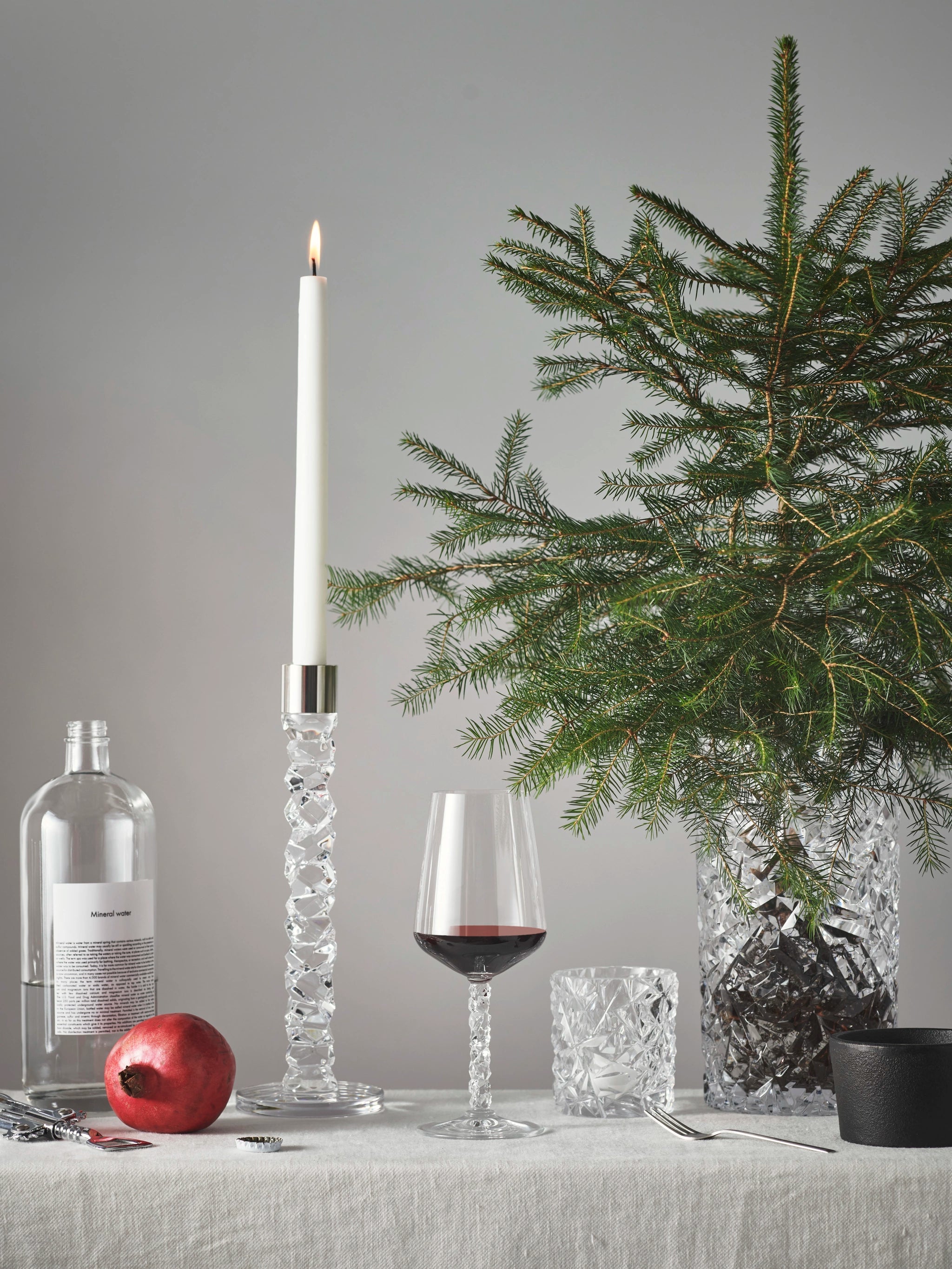 A lit candle, a 14.8oz Orrefors Carat wine glass filled with red, a pomegranate, an empty bottle, and a pine branch in a vase sit gracefully on the table with its light tablecloth.