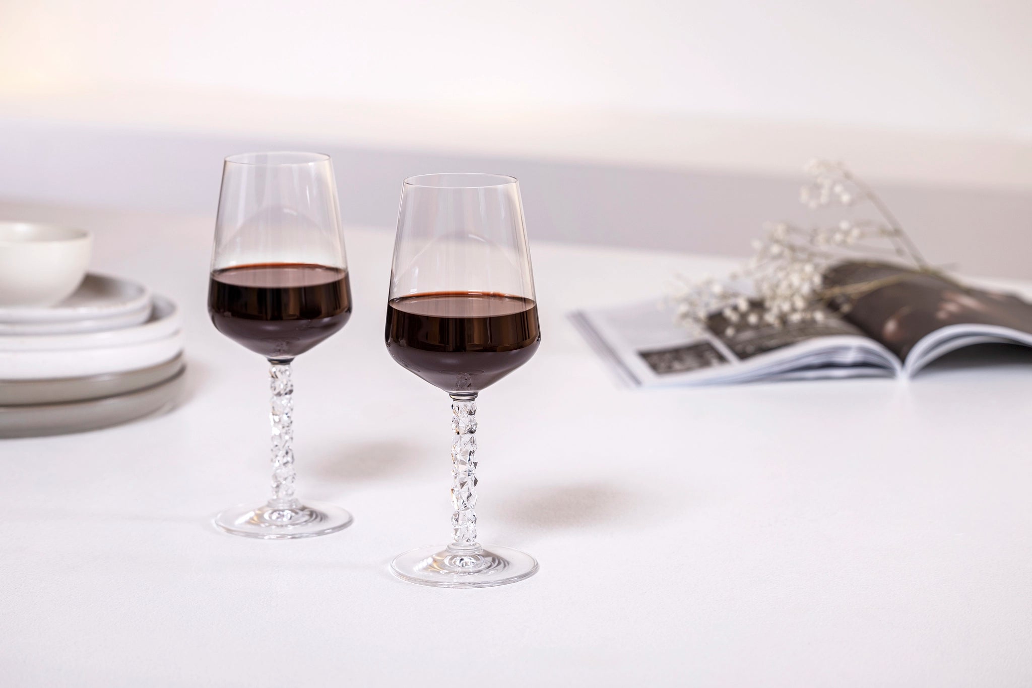 Orrefors Carat Wine Glasses, each holding 14.8oz of red wine, rest on a white surface with stacked plates and an open magazine decorated with flowers.