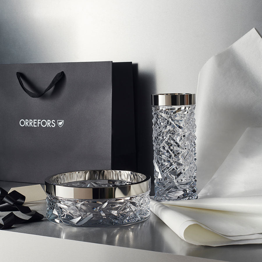 The Orrefors: Carat Bowl & Vase Bundle dazzles with metallic accents on a reflective surface and comes with an Orrefors gift bag, black ribbon, and white tissue paper.