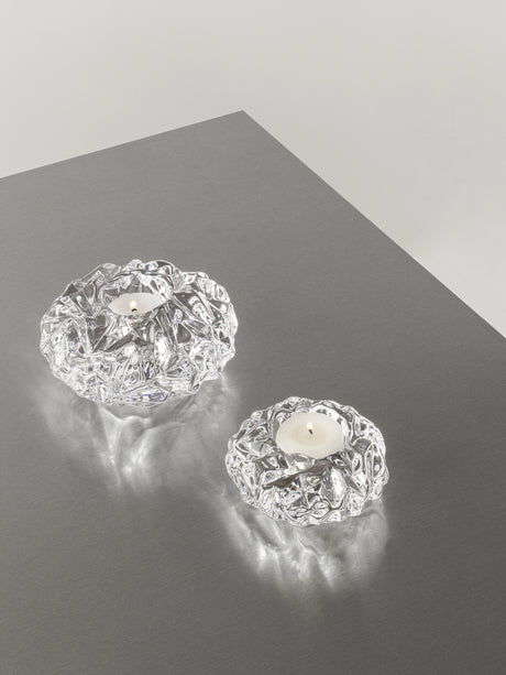 Two Orrefors: Carat Votive Small Candle Holders with intricate designs grace a gray surface, each holding a lit tealight and adding an elegant sparkle to any setting.