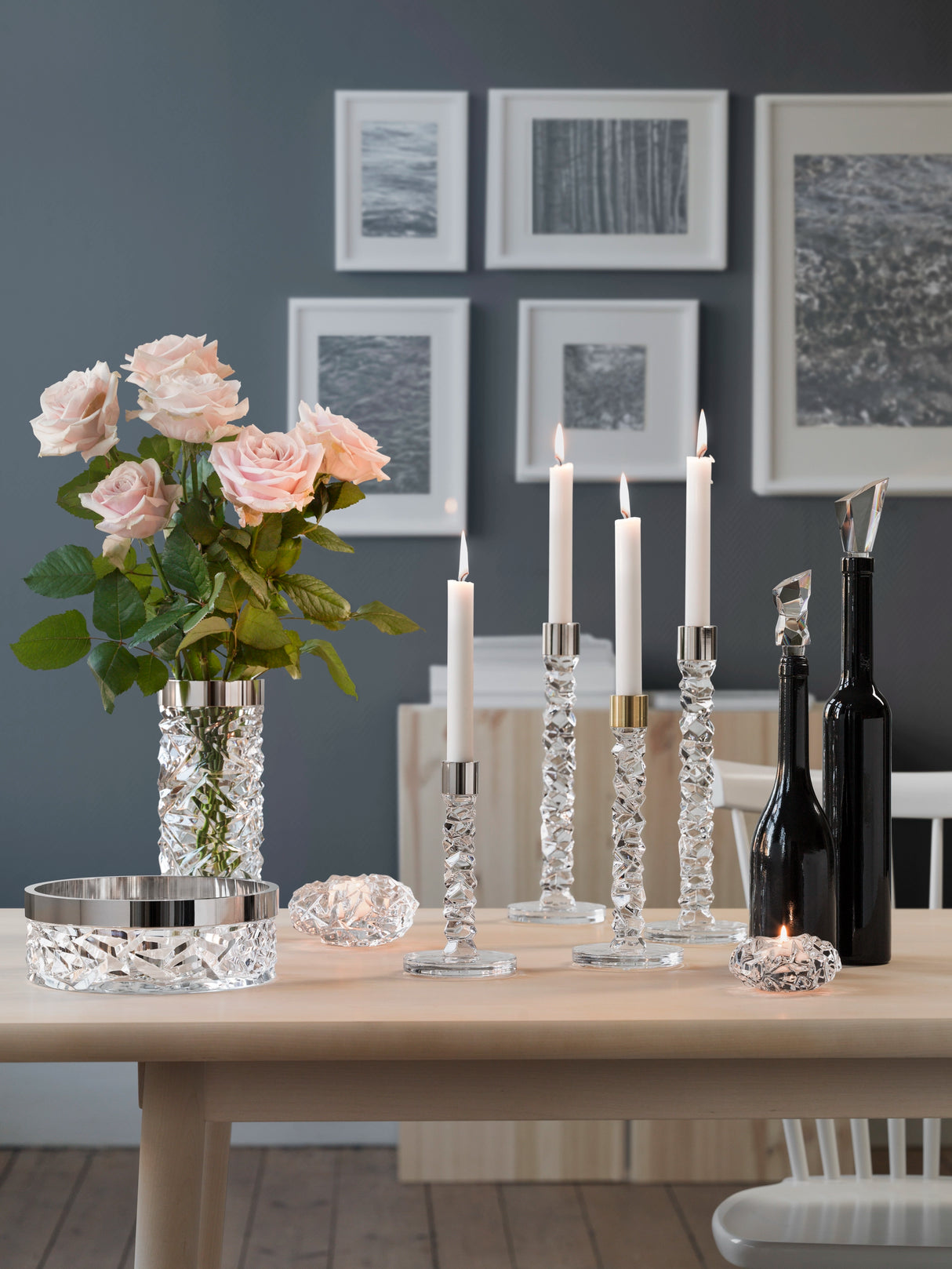 Elegant table setting featuring white candles in an Orrefors Carat Votive Small Candle Holder, crystal holders, a vase of pink roses, black decorative bottles, and framed art on a gray wall.