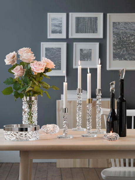 Elegant table setting featuring white candles in an Orrefors Carat Votive Small Candle Holder, crystal holders, a vase of pink roses, black decorative bottles, and framed art on a gray wall.