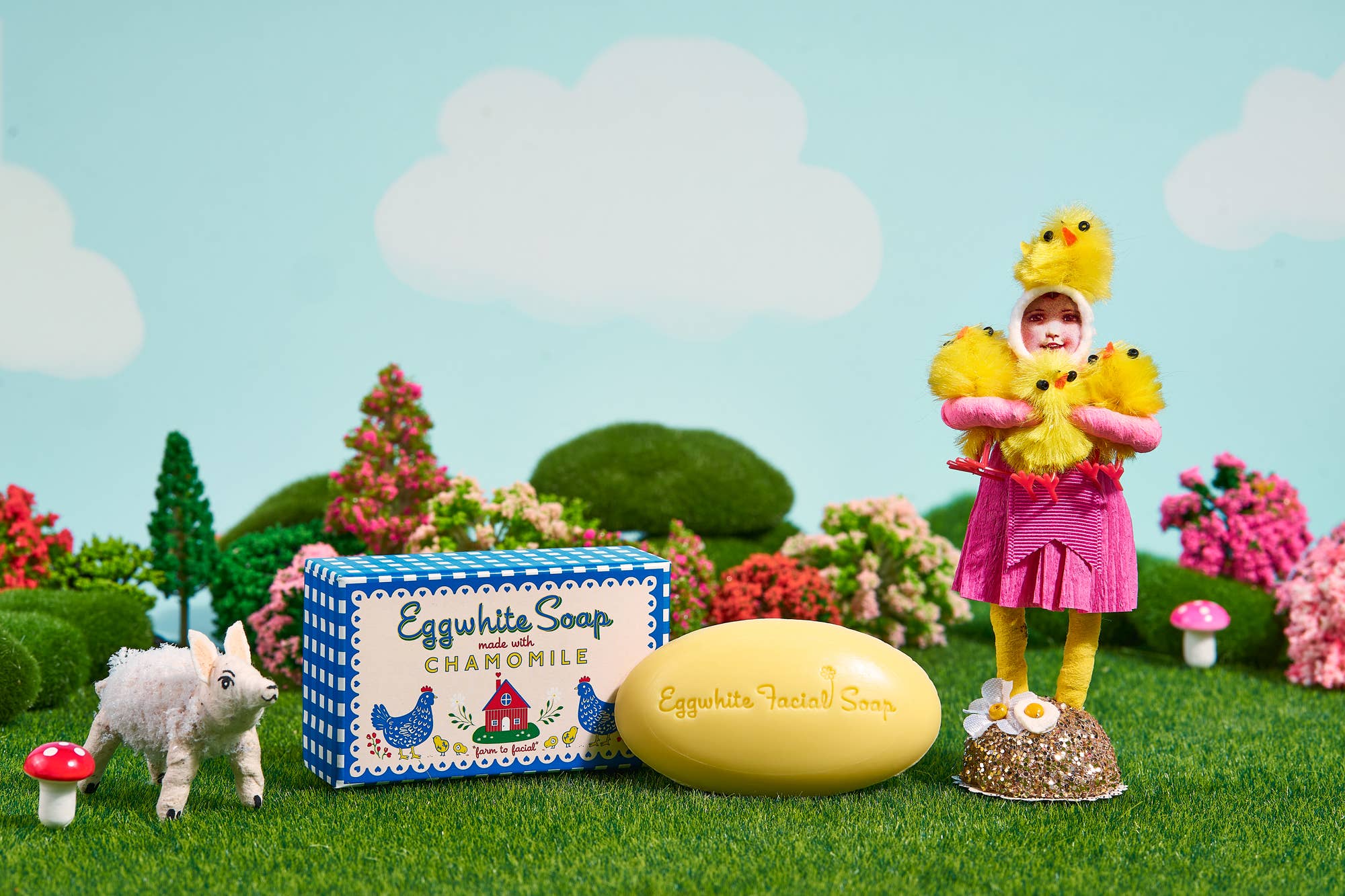 A child in a chick costume stands by a large Eggwhite & Chamomile Facial Soap in a gingham gift box, with a goat figurine and colorful scenery. This gentle soap helps tighten pores for fresh, radiant skin.
