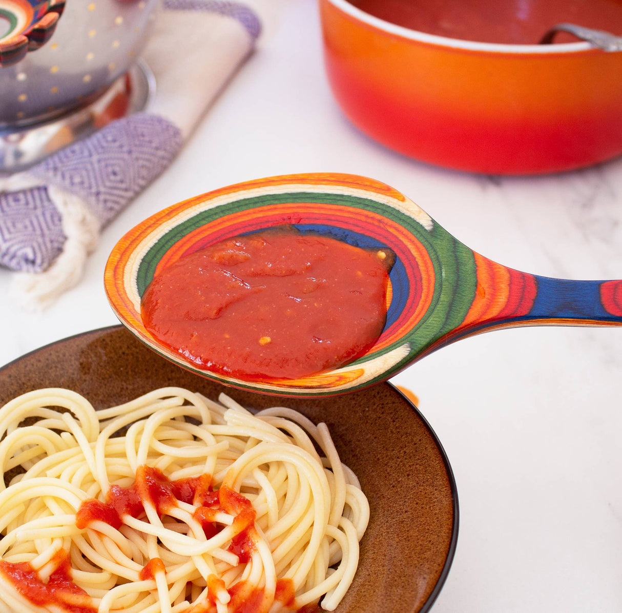 Serving spoon pouring tomato sauce over plain spaghetti, with a red pot and folded kitchen towel in the background on a white surface.