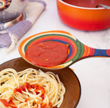 Serving spoon pouring tomato sauce over plain spaghetti, with a red pot and folded kitchen towel in the background on a white surface.