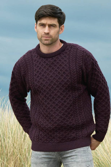 A man stands outdoors in front of tall grass and a cloudy sky, wearing light jeans and the Merino Crew Neck Sweater in Damson.