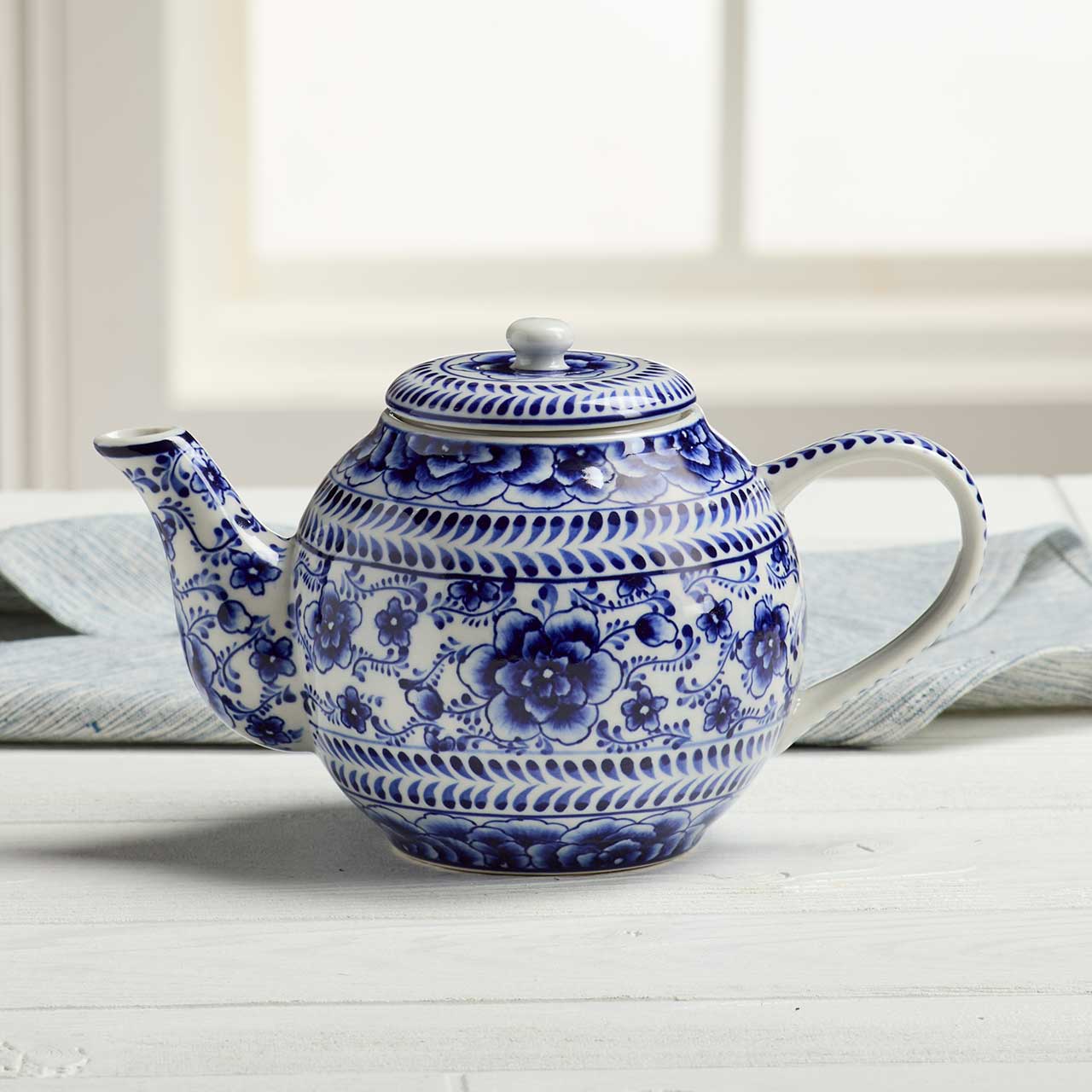 The Indigo Bloom Tea Infuser Pot, a blue and white floral ceramic teapot, sits on a white table with a folded cloth and window behind it, its hand-painted design bringing charm to any tea setting.