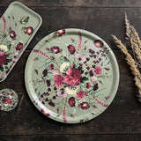Round Nordic birch veneer tray with red, pink, and white flowers and green leaves on a muted green background, placed on a dark wooden surface.