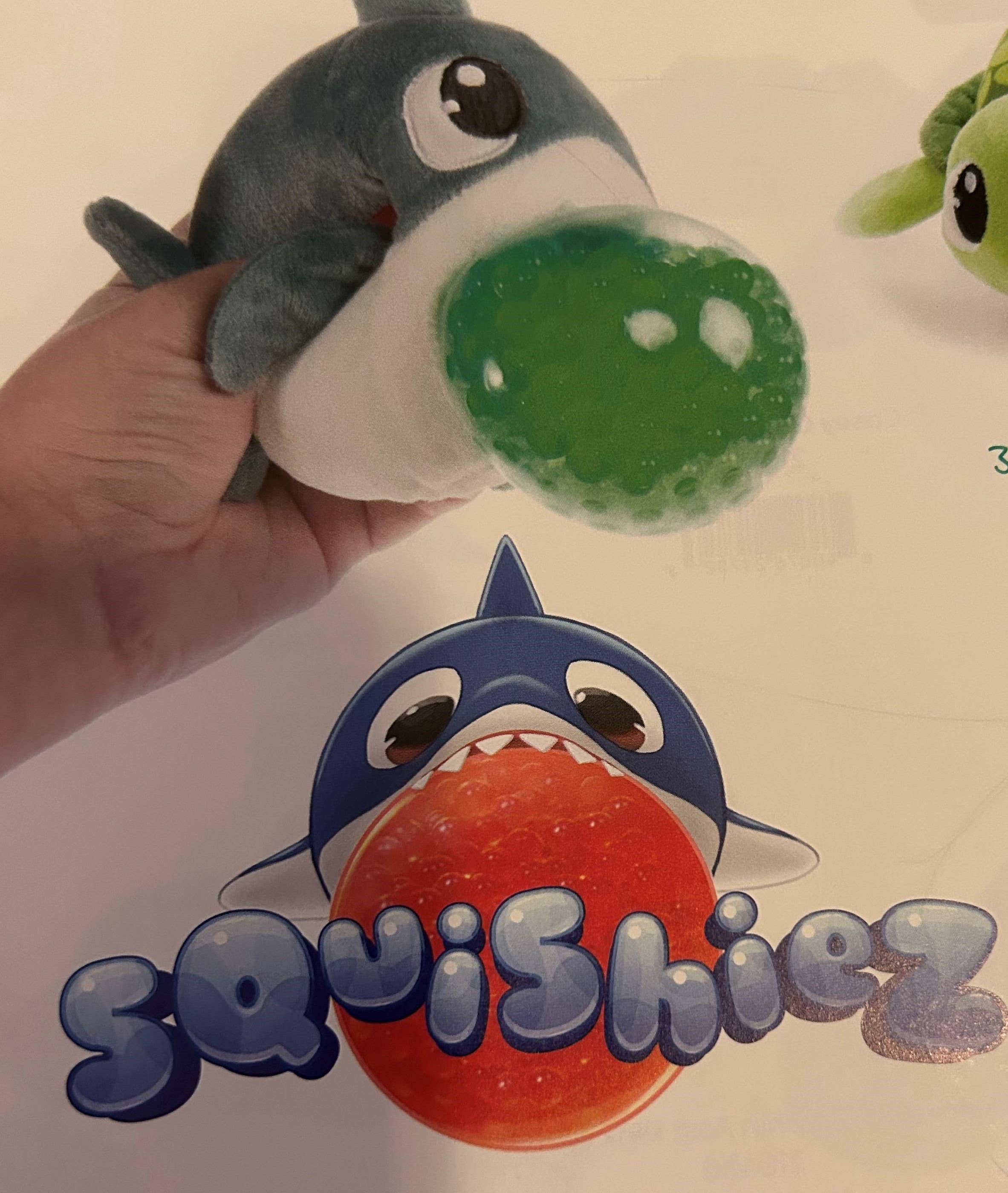 A hand holds a plush toy penguin, part of the Squishiez collection, featuring a green gel bead-filled ball popping from its mouth for squishable fun. Below, an illustration shows the same type of penguin with an orange ball and the text reads "Squishiez: Penguin Small 3".