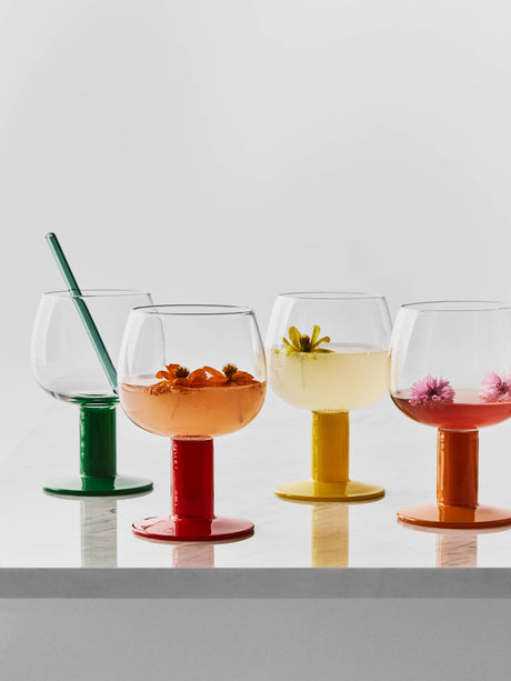 Four mouth-blown glass goblets with green, red, yellow, and orange stems, each filled with a beverage containing an edible flower and a green glass straw.