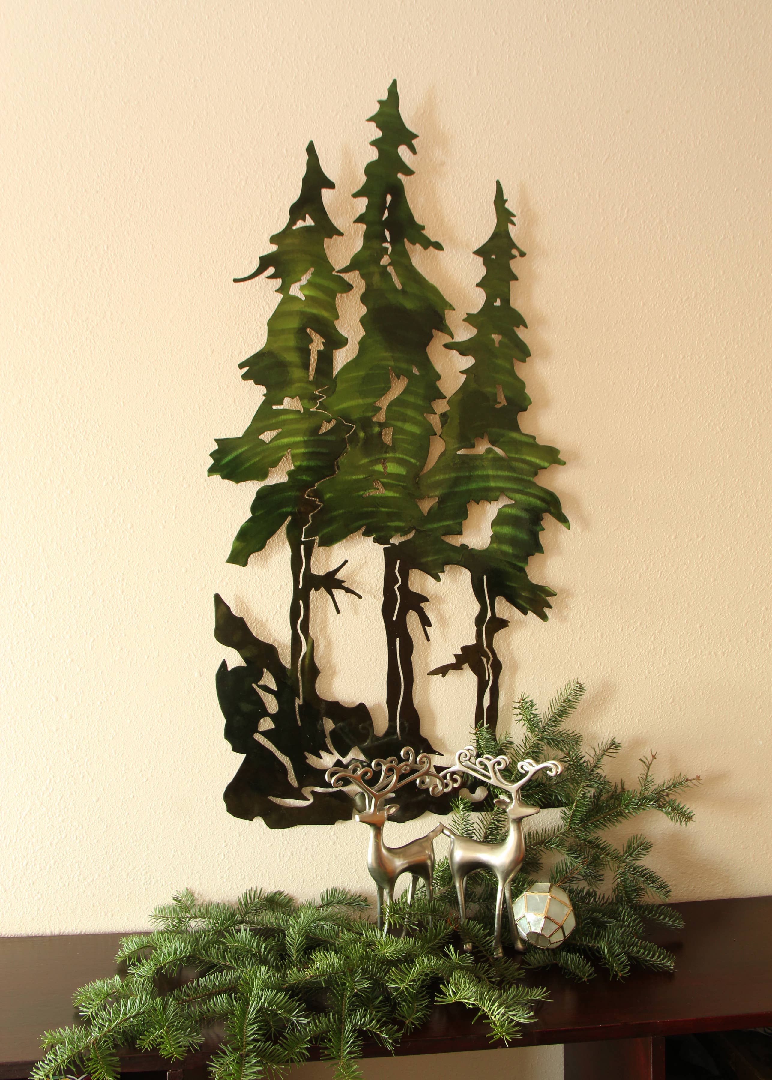 Wall Decor: Pine Tree Metal Wall Art: 22", featuring a sculpture of three pine trees and two decorative deer with evergreen branches, beautifully displayed on a shelf.