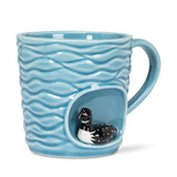 The Wavy Aqua Mug with Loon in Recess (4"H, 14oz) is a handcrafted blue ceramic mug featuring a wavy texture and a small black-and-white loon figurine at the base—an elegant, nature-inspired gift.