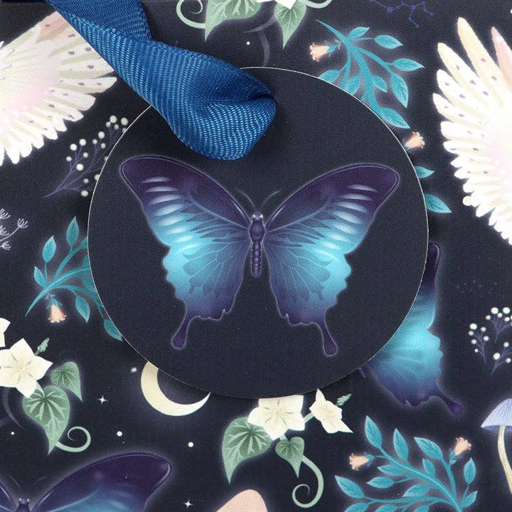A circular dark tag with a blue butterfly illustration hangs from a blue ribbon on the Midnight Forest Owl Print Medium Gift Bag, which features a dark floral and celestial-patterned background.