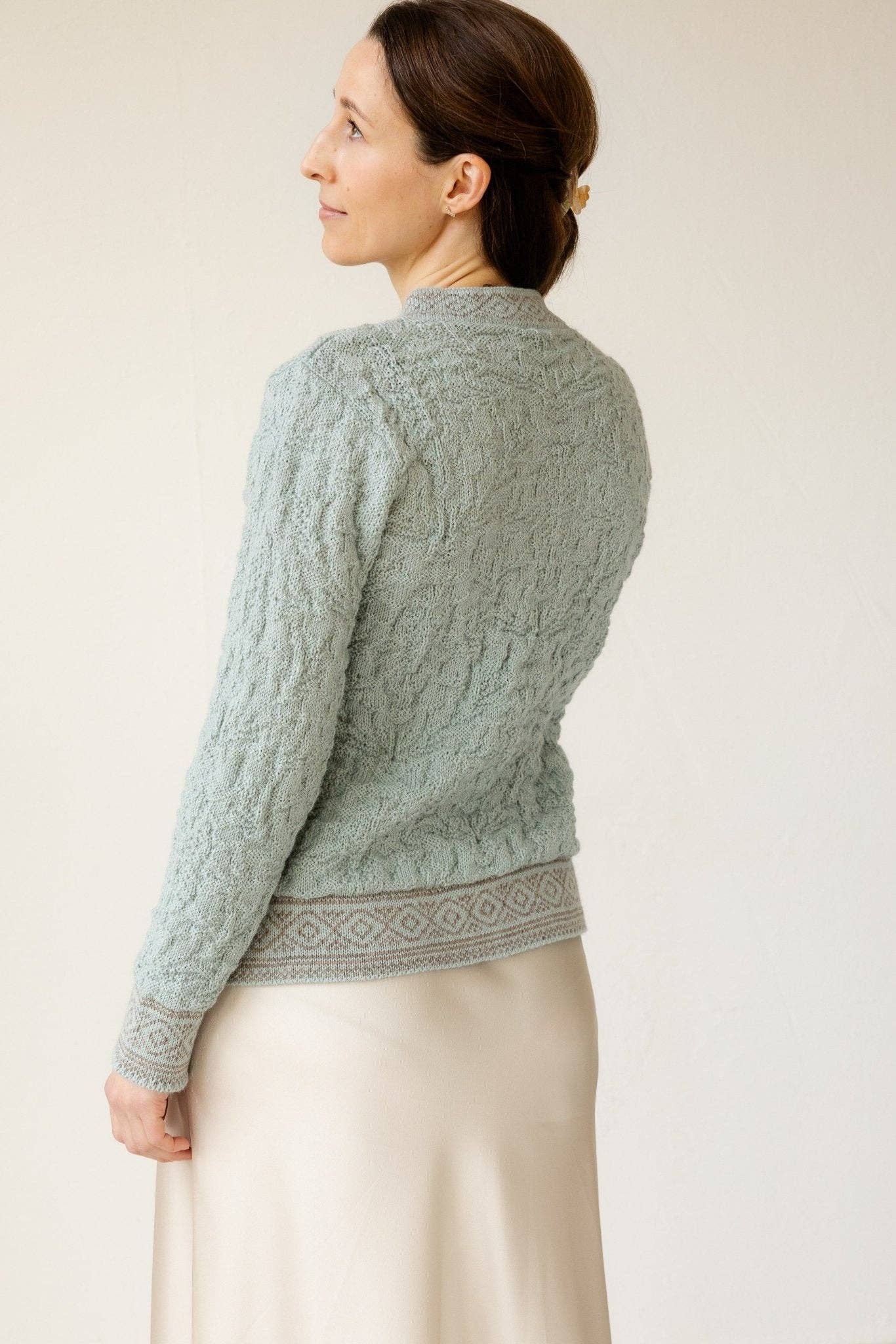 A woman stands sideways, wearing the Bryggen Women's Norwegian Cardigan (White)—crafted from 100% wool with textured patterns—and a cream skirt, gazing slightly upward.