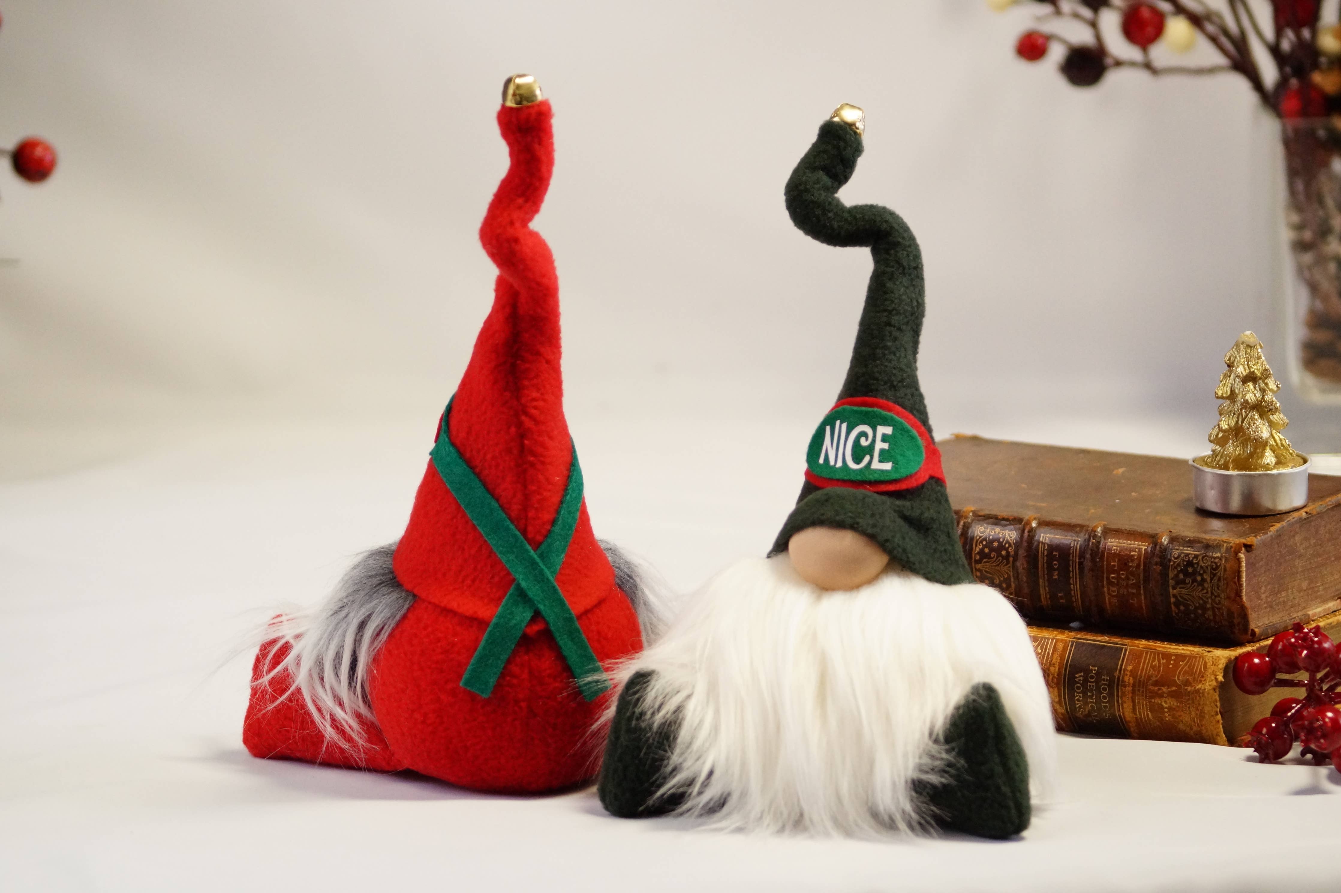 Two festive Nice Christmas gnomes with tall hats are placed on a table. One gnome features a red hat adorned with a green X, while the other showcases a green hat with NICE written on it. A small tree ornament and some books enhance the cozy scene featuring these charming, handmade gnomes.