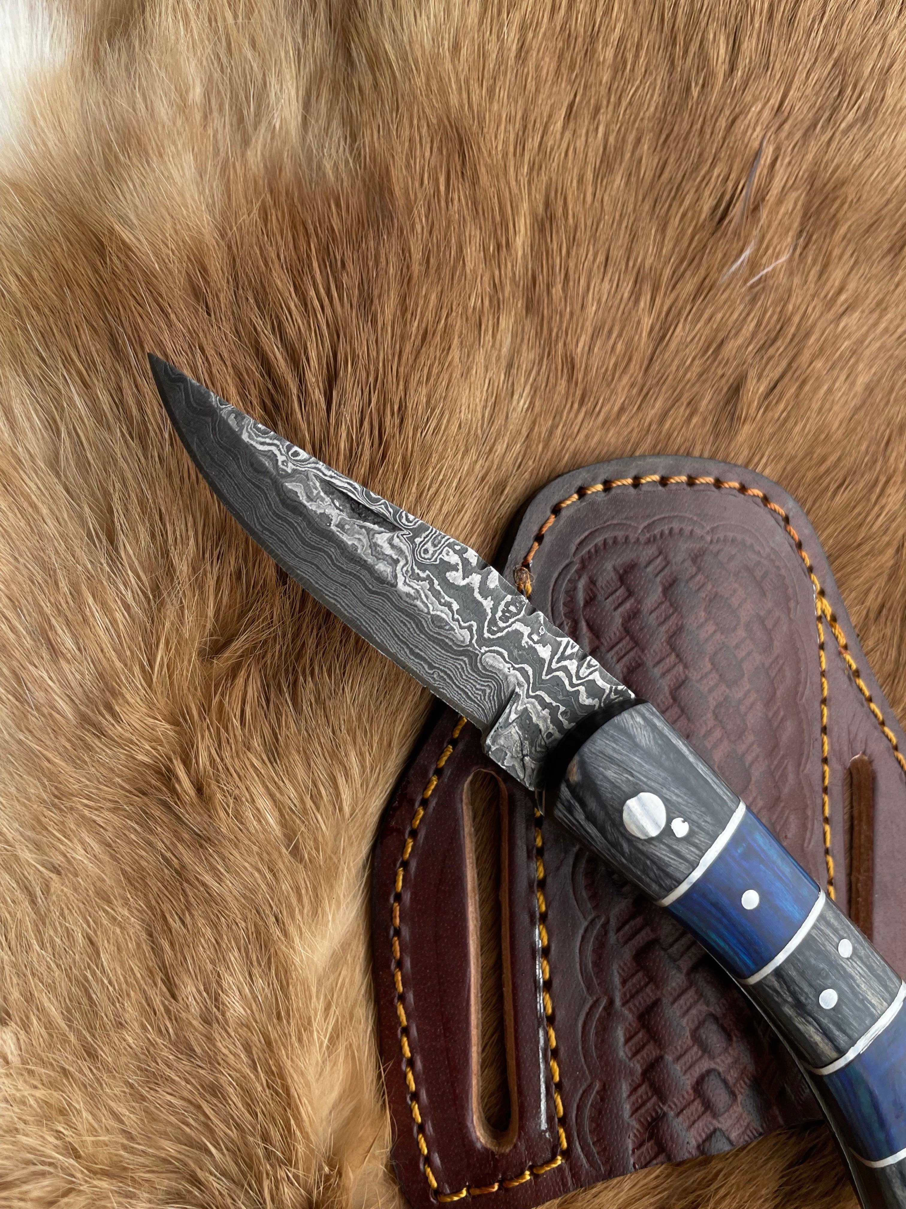 The Grey and Blue Wood 6.5 Damascus Steel Folding Knife, featuring a patterned blade and decorative handle, rests on a brown leather sheath atop animal fur.