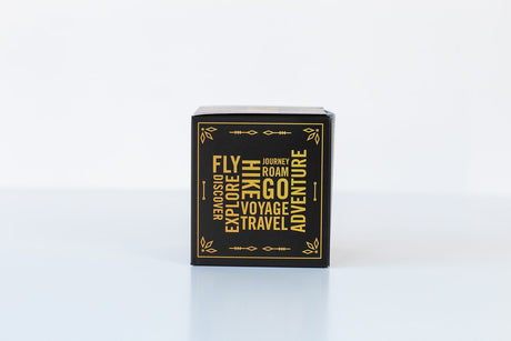 A small black box with gold lettering featuring travel-inspired words like FLY, EXPLORE, and ADVENTURE on the front, perfect for gifting the Soap: Sandalwood & Vanilla Bath Bomb.
