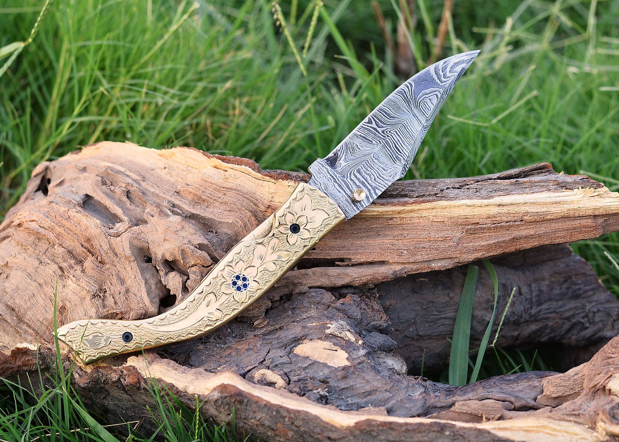 The Exquisite Golden Folding Knife, showcasing a patterned stainless steel blade and ornate handle, is elegantly displayed on driftwood with a grassy backdrop.