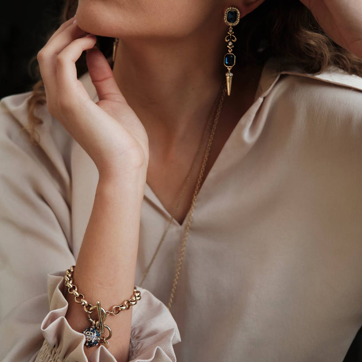 A person in a light beige blouse showcases a gold bracelet, a gold necklace, and A&C Oslo Antoinette Midnight Blue Statement Earrings—gold plated with midnight blue stones.