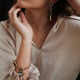 A person in a light beige blouse showcases a gold bracelet, a gold necklace, and A&C Oslo Antoinette Midnight Blue Statement Earrings—gold plated with midnight blue stones.
