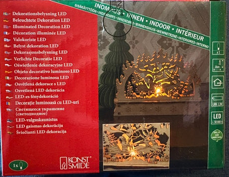 Glowing wooden LED decor with a winter scene placed before a fireplace; packaging displays text descriptions in several languages.