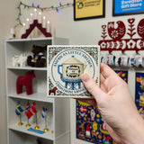 A hand holds a square coaster featuring a Lindström coffee-pot water tower illustration, floral accents, a Dala horse, and Swedish text.