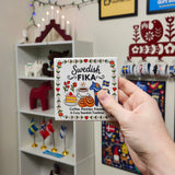 A hand holds a square Swedish Fika ceramic coaster featuring a Dala horse, coffee, pastries, and floral borders.