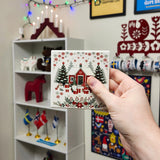 A hand holds a square Swedish folk art coaster featuring a red house, pine trees, and a tomte gnome surrounded by wrapped gifts.