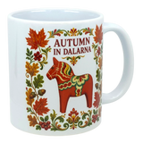 White mug featuring Scandinavian folk art with autumn leaves, acorns, a red Dala horse, floral patterns, and text reading “Autumn in Dalarna.”.