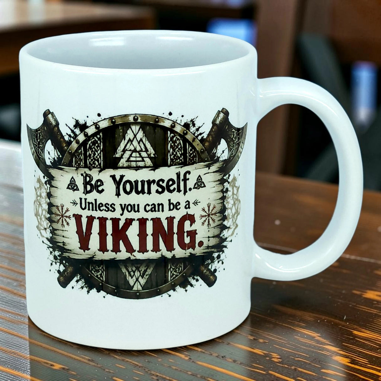 A white 11 oz ceramic mug from Nordic Wholesale, the "Be Yourself Unless You Can Be A Viking" Mug, features a bold design with a Viking shield and axes, and striking red "Viking" lettering. Ideal for fans of Norse mythology, this unique mug adds character to any wooden table.