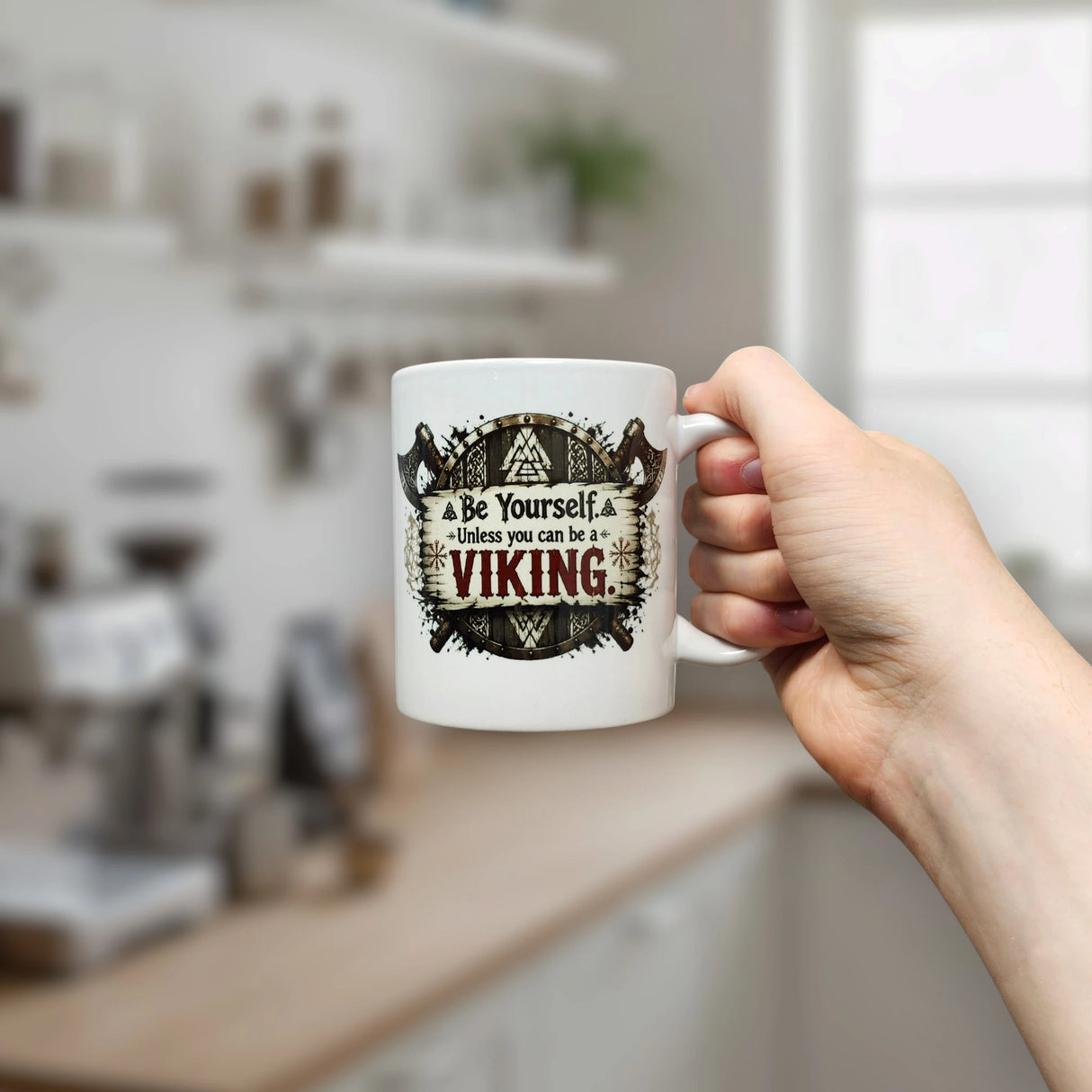 A hand holds the Nordic Wholesale "Be Yourself Unless You Can Be A Viking" ceramic mug (11 oz) in a kitchen. The white mug features playful text surrounded by a decorative Viking helmet and axes, reflecting the bold spirit of Norse mythology.