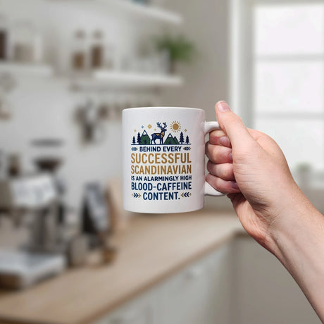 In a kitchen, a hand holds the Swedish Gift Store's 11 oz "Behind Every Successful Scandinavian Ceramic Mug," featuring the witty quote and playful designs of trees, a moose, house, and sun on its white surface.