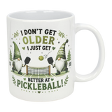 The Nordic Wholesale "Better at Pickleball" Ceramic Gnome Mug (11 oz) features two Nordic gnomes with paddles, net, and ball, green accents, and fun text: "I don't get older, I just get better at pickleball!" A perfect gift for pickleball fans.