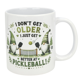 The Nordic Wholesale "Better at Pickleball" Ceramic Gnome Mug (11 oz) features two Nordic gnomes with paddles, net, and ball, green accents, and fun text: "I don't get older, I just get better at pickleball!" A perfect gift for pickleball fans.