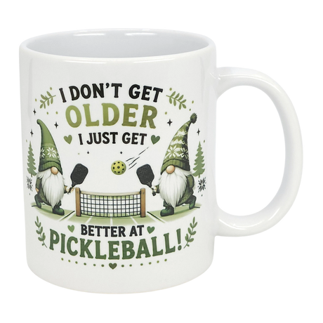 The Nordic Wholesale "Better at Pickleball" Ceramic Gnome Mug (11 oz) features two Nordic gnomes with paddles, net, and ball, green accents, and fun text: "I don't get older, I just get better at pickleball!" A perfect gift for pickleball fans.