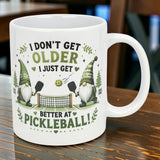 The Mug: Better at Pickleball Ceramic Gnome Mug (11 oz) by Nordic Wholesale features two Nordic gnomes with pickleball paddles beside a net and the phrase "I don't get older, I just get better at pickleball!" in playful text.