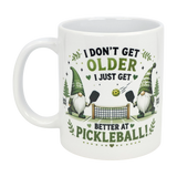 The Nordic Wholesale "Better at Pickleball" ceramic gnome mug (11 oz) features two Nordic gnomes playing pickleball, surrounded by leaves and hearts, with the message: I don't get older, I just get better at pickleball!.