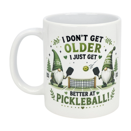 The Nordic Wholesale "Better at Pickleball" ceramic gnome mug (11 oz) features two Nordic gnomes playing pickleball, surrounded by leaves and hearts, with the message: I don't get older, I just get better at pickleball!.