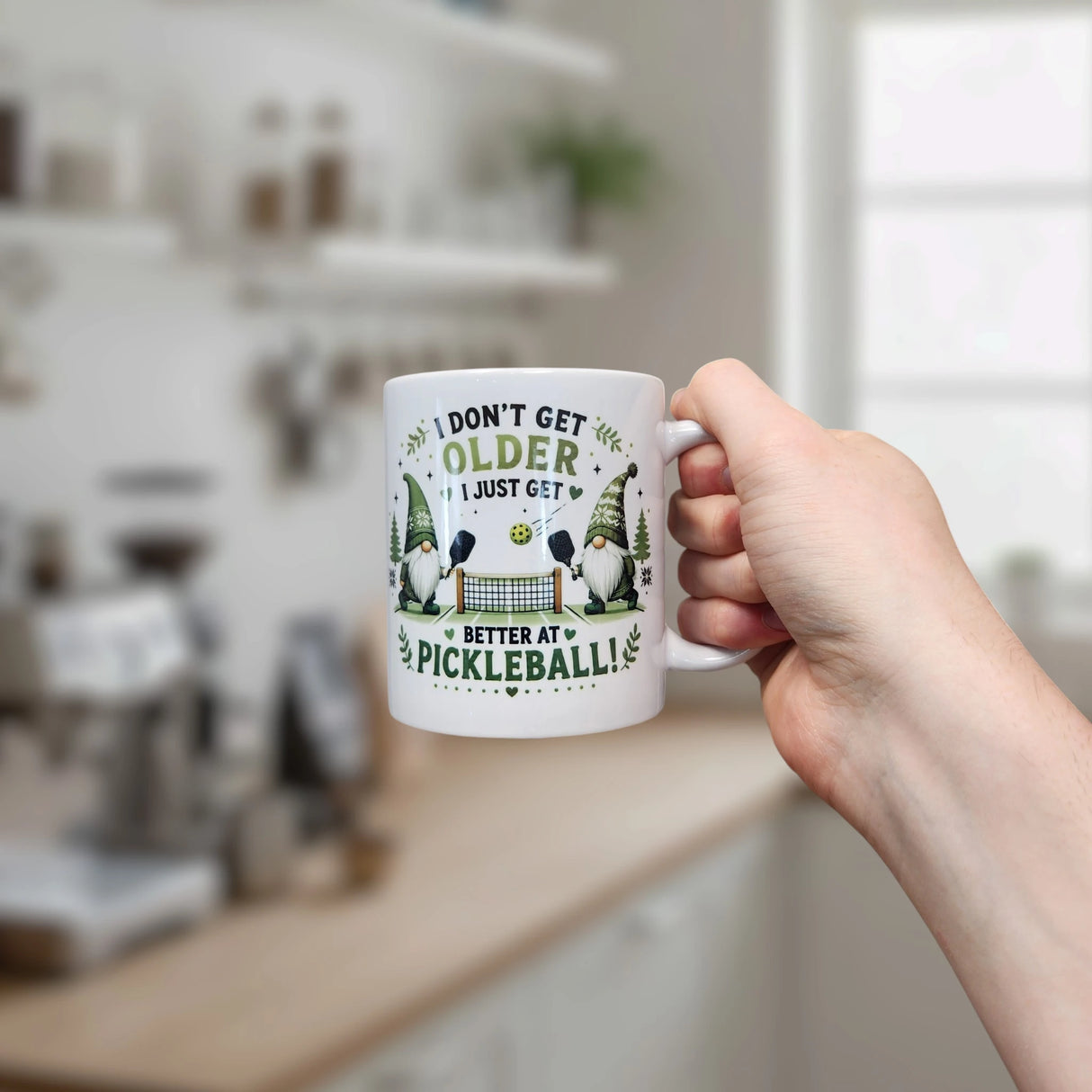 A hand holds the Nordic Wholesale Mug: Better at Pickleball Ceramic Gnome Mug (11 oz) in a cozy kitchen, featuring paddles and the phrase "I don’t get older, I just get better at pickleball" against a softly blurred background.