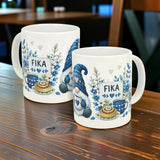 Two Nordic Wholesale Blue Tomte Gnome Fika Ceramic Mugs (11 oz) with blue and gray Scandinavian gnome and foliage designs, plus "FIKA" text, are displayed on a wooden table in a cozy setting.