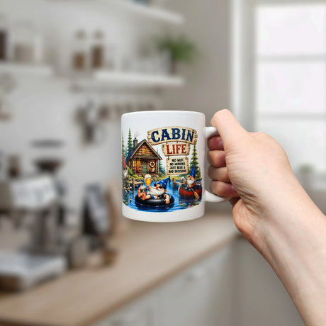 A hand holds the Nordic Wholesale Mug: Cabin Life Gnomes Ceramic Coffee Mug (11 oz) in a kitchen. The mug showcases artwork of a cabin, gnomes lounging on an inner tube with bears, trees, and the phrase “Cabin Life—No WiFi, just an improved connection.”.