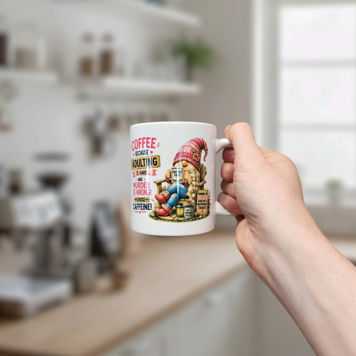 A hand holds the Nordic Wholesale Mug: Choose Caffeine Tomte Ceramic Mug (11 oz), showcasing a whimsical gnome and caffeine-inspired text, with a bright, blurred kitchen in the background.