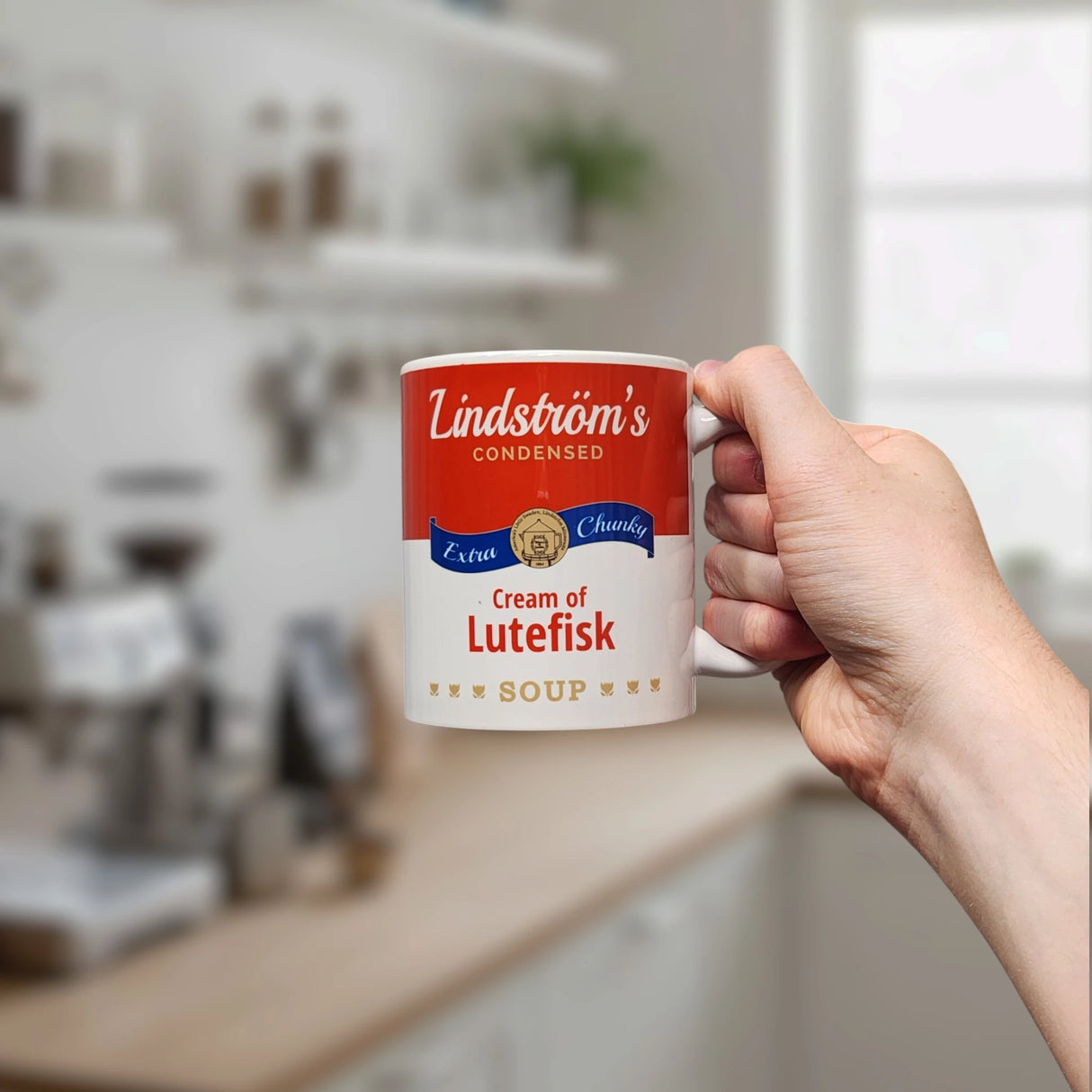 A hand holds the Swedish Gift Store’s 11oz ceramic mug, designed like a soup can label reading “Lindstrom’s Condensed Extra Chunky Cream of Lutefisk Soup,” with a blurred kitchen backdrop—an ideal Scandinavian heritage gift.