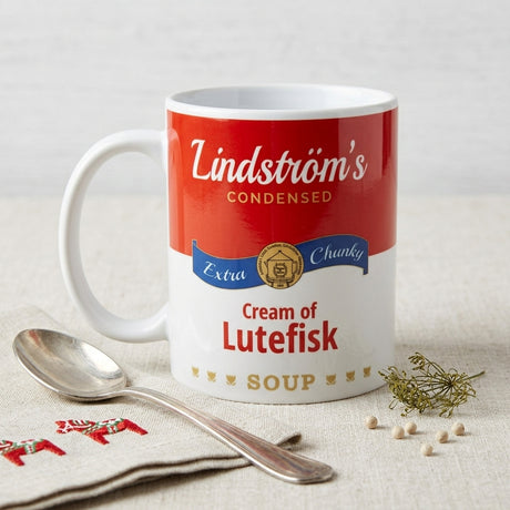 A mug designed like a soup can labeled Lindströms Cream of Lutefisk Soup on a cloth with a spoon, fresh dill, and an embroidered napkin.