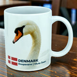 Introducing the Mug: Denmark National Animal Mute Swan Ceramic Mug (11 oz) by Nordic Wholesale. This white ceramic mug features a beautiful image of the graceful Mute Swan, accompanied by the Danish flag and the words "DENMARK Knopsvane | Mute Swan." Perfect for adding a Scandinavian touch to your kitchen, this Danish flag mug from Nordic Wholesale celebrates iconic Nordic elegance and is beautifully displayed on a wooden table.