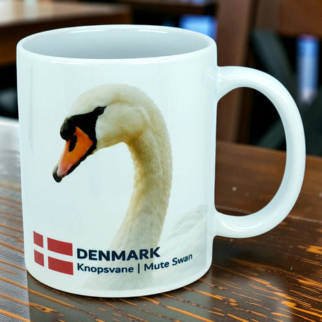 Introducing the Mug: Denmark National Animal Mute Swan Ceramic Mug (11 oz) by Nordic Wholesale. This white ceramic mug features a beautiful image of the graceful Mute Swan, accompanied by the Danish flag and the words "DENMARK Knopsvane | Mute Swan." Perfect for adding a Scandinavian touch to your kitchen, this Danish flag mug from Nordic Wholesale celebrates iconic Nordic elegance and is beautifully displayed on a wooden table.